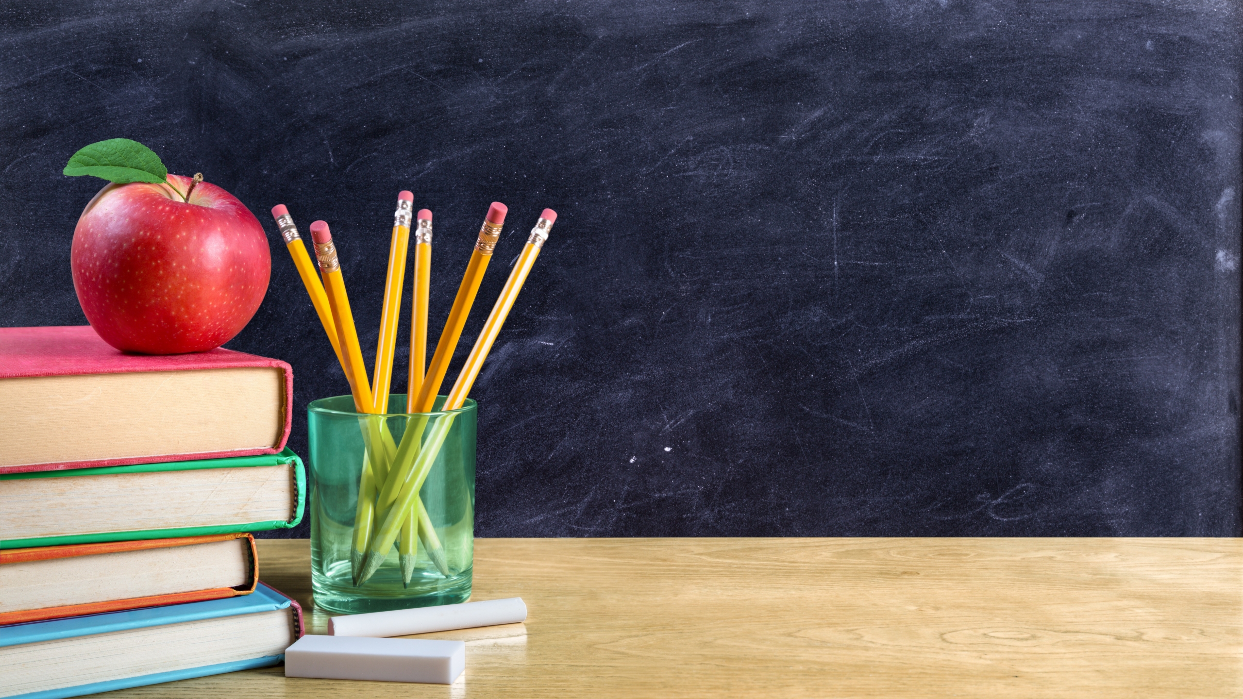 Stacked books, a red apple, and a glass of yellow pencils in front of a black chalkboard.