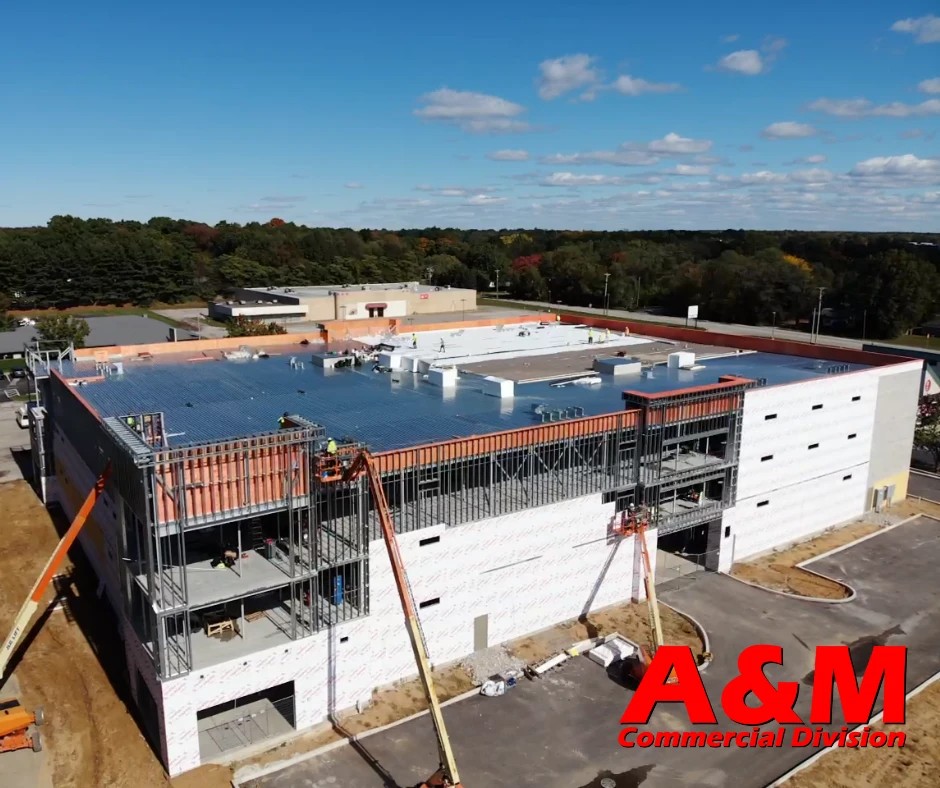 An aerial view shows multiple boom lifts stationed around a large commercial facility under construction as workers install a massive flat roof system, demonstrating the scale of professional commercial roofing services.