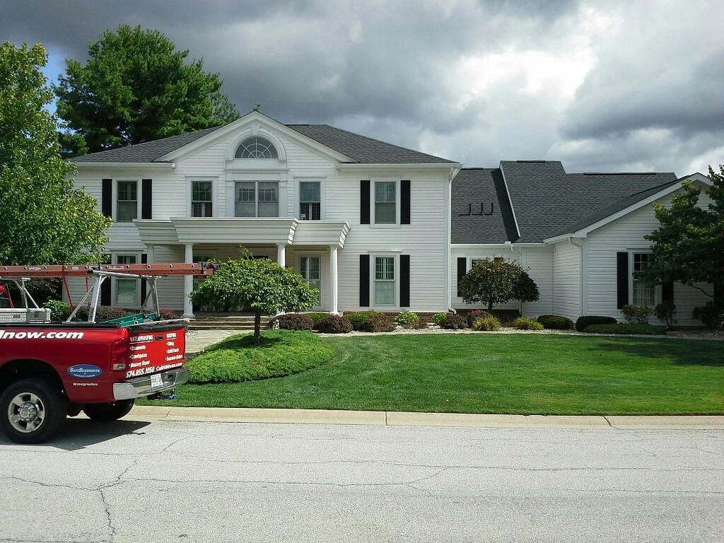 A red service truck is parked along the street in front of a sprawling white two-story home with dark shutters, highlighting a property upgraded by professional exterior services.