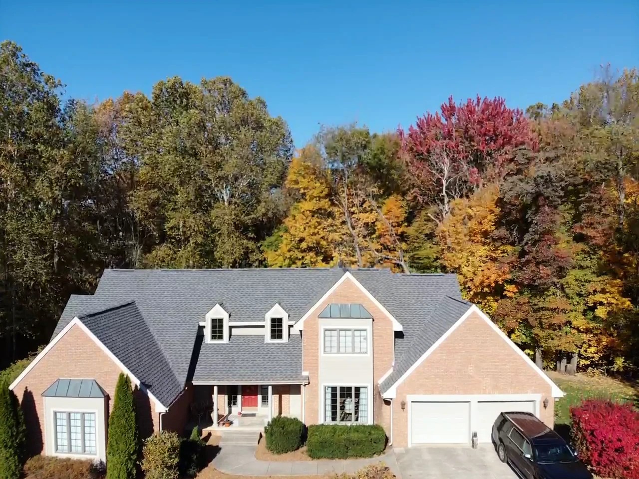 A two-story brick residence with dormer windows and a clean grey shingle roof sits before autumn foliage updated through complete exterior services.
