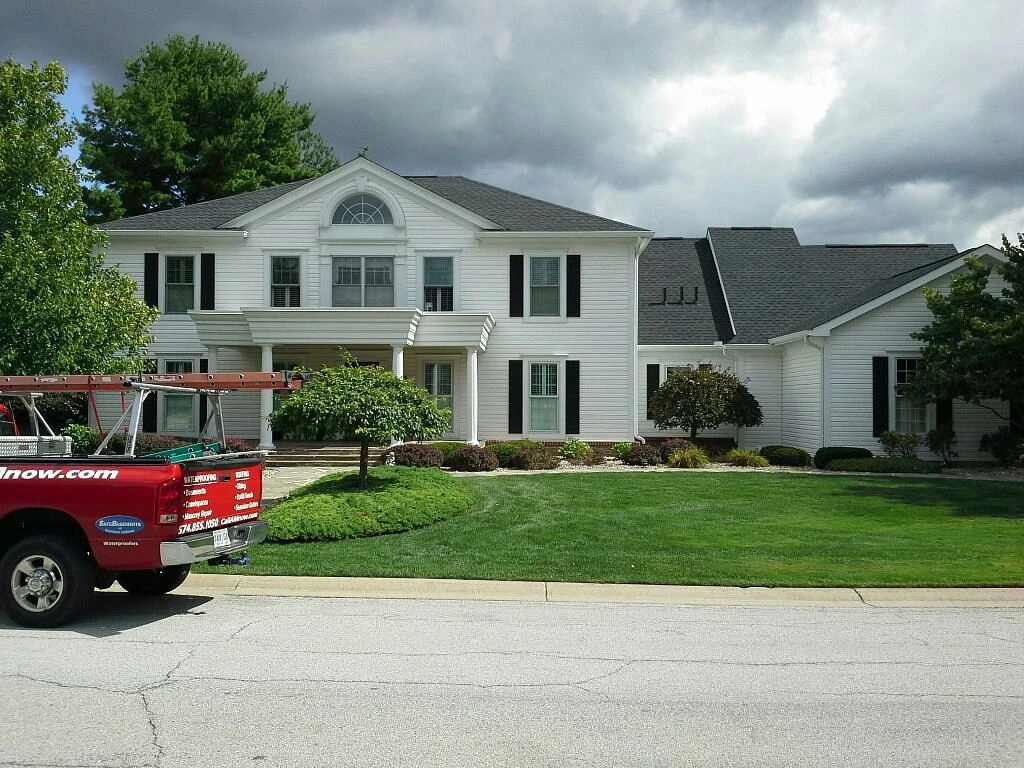 A red service truck sits parked in front of a grand two-story white home with completed dark shingle roofing services.